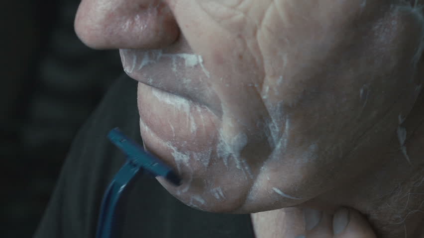 Close up profile of old gray man shaving beard on face by himself