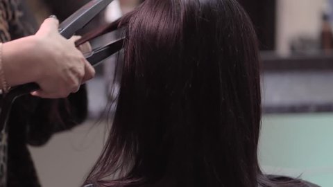 Hairstylist straightening the long black hair of a female client using a heated hair straightener. Slow motion. Close up.