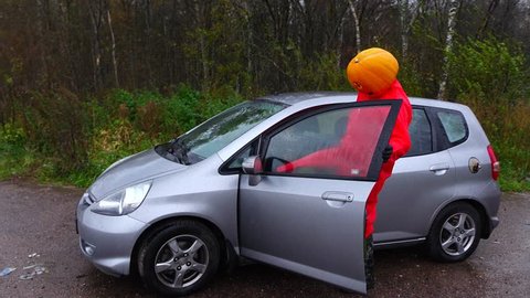 Man wear weird foolery costume, Halloween pumpkin on head, red rainsuit, come out small car, walk to road. Rainy outdoors at autumn season. Prank at rural area, freak guy have carved squash plant mask