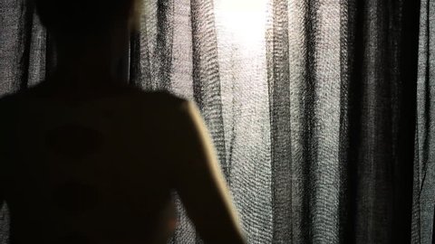 Young woman unveiling curtain, looking out of the window and enjoying beautiful day light. Beautiful image of the back of young girl with lens flare