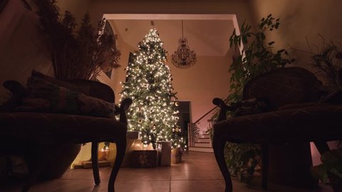 Big Christmas tree with glowing lights, ornaments, and presents in a beautifully decorated home. Dolly shot.