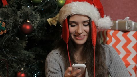 Pretty caucasian woman calling on the phone near the christmas tree. Attractive brunett woman in santa's hat greeting somebody with Christmas. Beautiful young woman holding cellphone near her ear