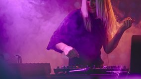 Tilt up of seductive female DJ with blond hair dancing behind decks and singing at party in nightclub