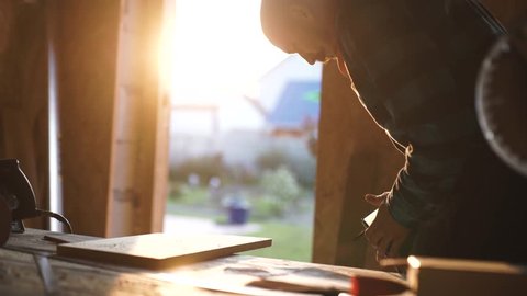 young bold carpenter with moustache. circular saw cutting piece of wood. sun flare on background