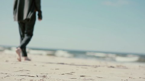 A woman with long dark hair is walking barefoot on a sandy beach towards the waves.