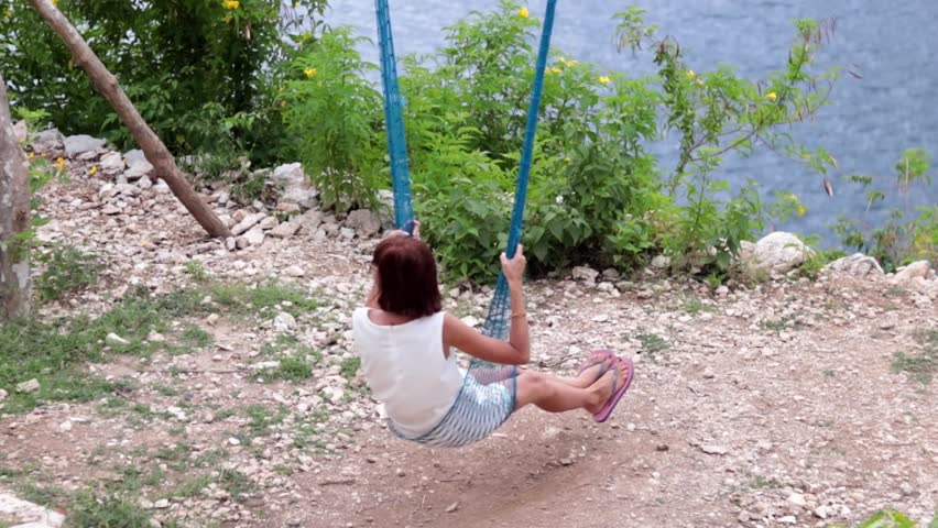 Young girl swinging on a swing in the mountains on a cliff above the ocean. Fantastic view, slow motion. Tropical island Ceningan, Indonesia.