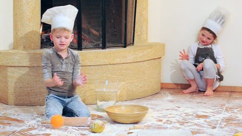 Kids in chef's hats near the fireplace: pretty 6 years girl is playing cook with her blue eyed cousin, they’re totally stained with flour