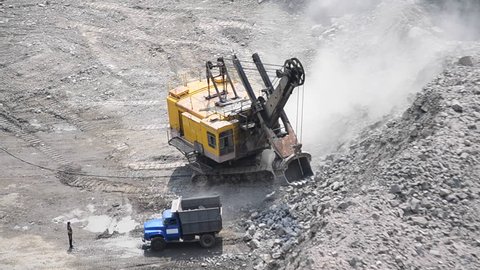 big huge yellow excavator moving on wheels tracks and revolves around its axis in a granite quarry and a man standing next to the waiting on a sunny day, lot of dust, top view with sound hd 1080