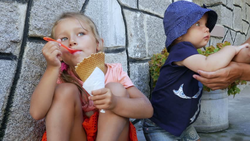 Two children eating ice cream on summer street, mom feeding baby girl ice cream, funny faces of kids