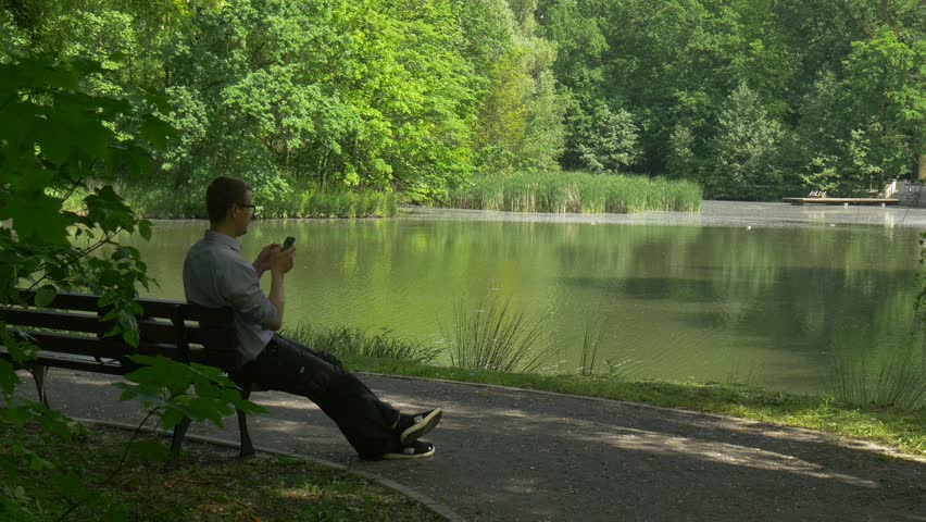Tourist is Resting on a Bench, Then Makes a Call on Mobile Phone in Park, Sitting on a Bench and Talking, Backpacker in Glasses Leans Forward, Stretched His Legs, Young Man Near the Lake in Park or