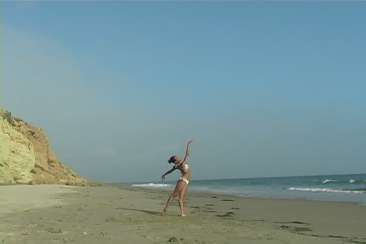 A beautiful young woman performs on the beach as horses pass by.   