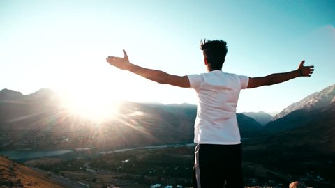 young Asian male reaching to the top of the mountain area, standing on top of a mountain, hands raised, awareness of success, slow motion