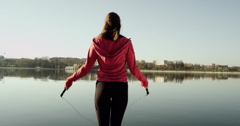 Close-up of back an jumping women on the jump rope. Outdoor sports. Girl jumping on a skipping rope near lake. City and park on background. 4K 