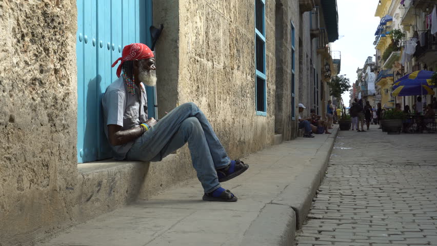 HAVANA, CUBA - SEPTEMBER 15, 2016: Elderly Afro-American man with gray-haired beard and braids in red bandanna sits on step near a house, have crossing the legs and looking street at sunny summer day