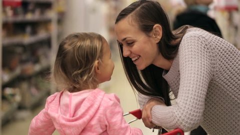 Smiling young mother and cute little daughter talking at the supermarket. girl sitting in trolley, smiling and hugs her mom.