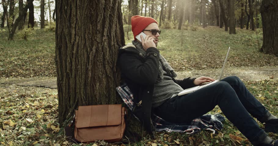 A young man in a park using a laptop and a mobile phone