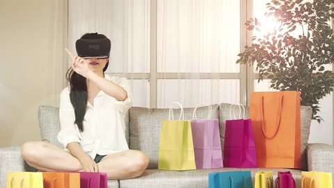 asian woman experience shopping online with VR headset sitting on sofa colorful shopping bags around. indoor living room background