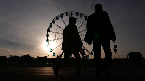 Two citizens walk by and come away against Ferris Wheel, silhouetted view. High contrast scene of Helsinki SkyWheel at sunny evening, bright sun beam and shaded skies, black construction contour Stock Video