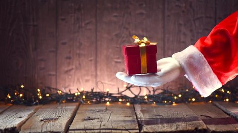 Santa Claus holding gift box, gives a gift, holding it in his hand over wooden background with blinking Christmas tree garland. Full HD 1080p video footage