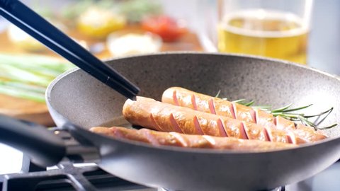 In the kitchen a woman is cooking bratwurst in a frying pan using the tongs during the day. Warm and tasty mood. 