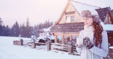 Woman with Cup of Hot Tea or Coffee by the Cozy Mountain Cottage on Snowy Winter. 4K SLOW MOTION 120fps. Beautiful Girl Enjoying Winter Outdoors, drinking from a Mug, getting warm. Christmas Holidays