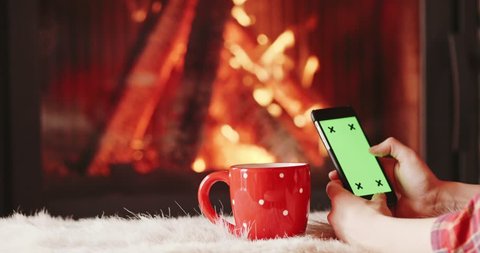Unrecognizable Woman Hands Using SmartPhone by the Burning Fireplace - Close Up. 4K SLOW MOTION 120FPS. Female hands with phone green screen and cup by cozy fireside. Searching internet, using app.
