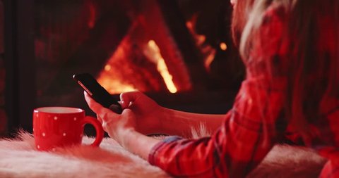Unrecognizable Woman Hands Using SmartPhone by the Burning Fireplace - Close Up. 4K SLOW MOTION 120FPS. Female hands with phone and cup of hot drink by cozy fireside. Searching internet, using app.