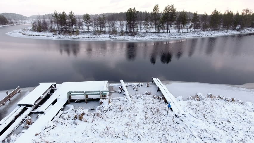 Drone flying up over snow covered timber pier on wintry river with open water. Winter river bend is in swamp. Karelia, Russia