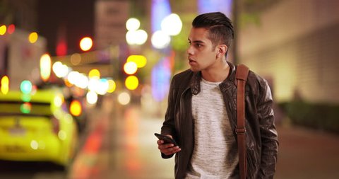 Millennial Latino man calling for taxi or ride sharing service on city street. Hispanic guy at night waiting for uber car with smart phone in hand. 4k