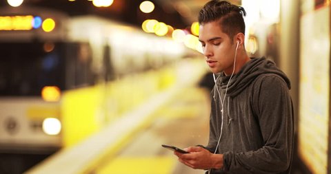 Millennial Latino man using smartphone waiting for subway train at metro station. Hispanic commuter using cell phone to listen to music at rail stop. 4k