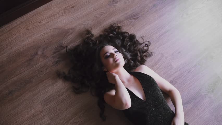 Sexy caucasian woman is laying on the floor in evening dress, beautiful girl with long hair in teasing dress, pretty woman holds her hand over the finery