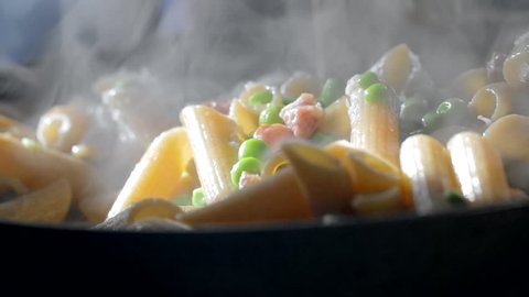 A woman is cooking penne pasta in a skillet dressed with green beans and diced bacon. Closeup slow motion shot.
