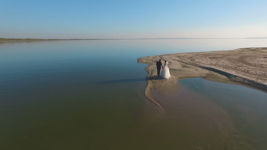 Attractive newly married couple taking time to themselves walking along a beautiful sandy beach. Aerial shooting.