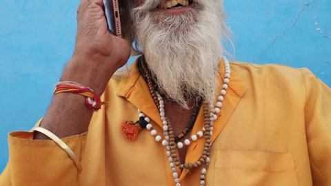 Tilt up closeup of a bearded Hindu priest wearing traditional saffron clothes on a call in Pushkar, Rajasthan