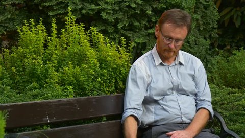 an Elderly Fair-Haired Man Looks Short Interesting Video on Youtube. he Does Not Take Away a Sight From a Screen of His Tablet Computer. the Man Has Conveniently Settled Down on a Wooden Bench in a