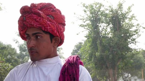 Indian man wearing a red turban talking to someone and then looks straight at the camera 