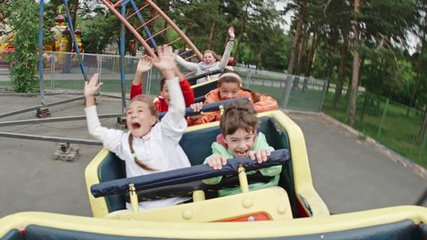 Joyful kids screaming and raising arms while riding roller coaster in amusement park