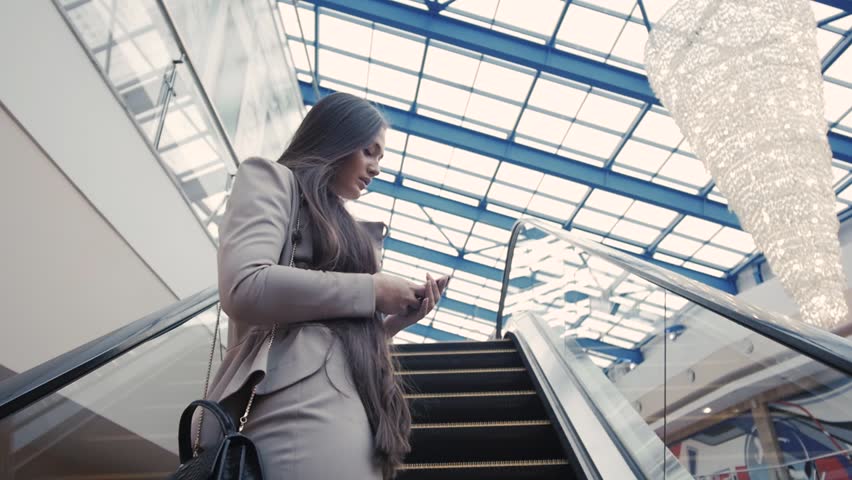 Confident woman is reading e-mail in internet via mobile phone, while is standing in shopping center on escalator. Female is booking on-line car, while is standing in airport near moving staircase Royalty-Free Stock Footage #21655141