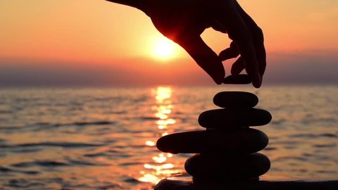 Cinemagraph loop background, balance concept. Beach scene in the cinemagraph technics: a man build zen stone pyramide at orange sea sunset background. Sea surf, wave, sun road. Cinemagraph HD footage.