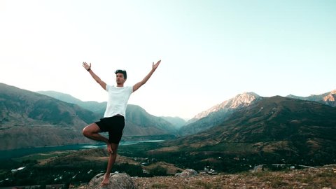 young Asian man doing yoga in a mountain area, standing on top of a mountain, healthy life, sun glare, slow motion