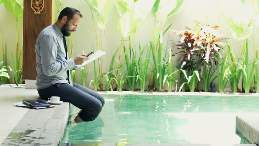 Young businessman working with smartphone and documents by pool in outdoor villa
