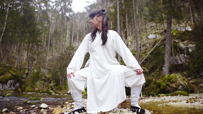 Woman Kung Fu person exercise dragon pose outside 4K. Wide shot sliding low angle view with person in focus making form above shallow river. Forest in background blur.
