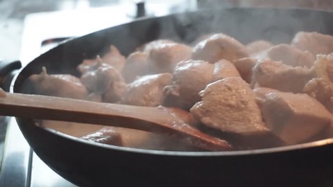 Concept: homemade, cooking, family. Close up of a chicken meat cooking in the frying pan. Woman hand is stirring it with a wooden turner. Slow motion 60fps