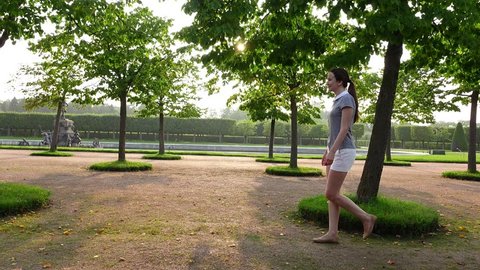 Tracking shot of positive girl quickly walking at Upper Garden of Peterhof park. Beautiful greenery, sunbeam flash through light green leaves of trimmed trees. Clean and accurate parkland