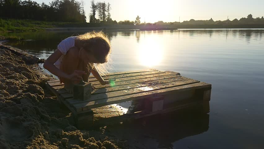Girl Sitting on the Haunches Near the Lake. Little Girl Playing With Sand and Stones Near the Lake. 