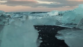 Dolly shot of ocean waves and icebergs on black volcanic sand beach during sunset
