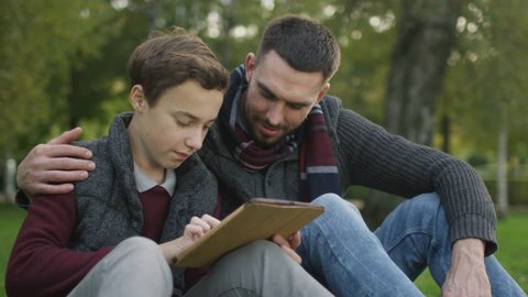 Son and Father Using Tablet Sitting in the Park. Shot on RED Cinema Camera in 4K (UHD).