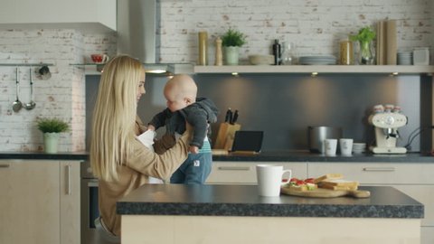 Young Mother Nursing Her Laughing Child while Standing on the Kitchen. Shot on RED Cinema Camera in 4K (UHD).