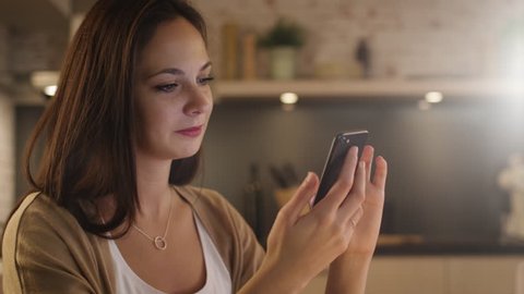 Beautiful Girl Uses Smartphone while Sitting in the Kitchen. She is Smiling. Shot on RED Cinema Camera in 4K (UHD).