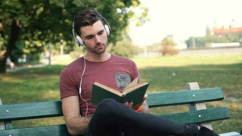 Handsome man listening music and reading book while sitting in the park
